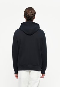 Hombre visto desde atrás con una sudadera con capucha negra y pantalones blancos frente a un fondo blanco liso.