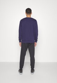 Marinblå sweatshirts med ribbad mudd och nederkant, kombinerad med svarta träningsoverallsbyxor. Modell sedd bakifrån, bärande mörka sneakers. Enfärgad bakgrund.