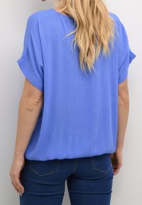 Blouse bleue légère à manches courtes, coupe ample, tissu texturé et col rond, associée à un jean en denim foncé.