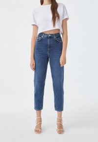 Jean en denim bleu taille haute avec une coupe slim, légère décoloration et cinq poches, associé à un t-shirt blanc court et des sandales blanches à lanières.