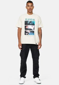 Beige grafisk t-shirt med dristige flerfarvede prints, der viser anime-ansigtter. Matchet med sorte cargo-bukser og hvide sneakers.