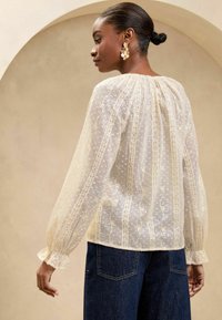 Translucent ivory blouse with textured fabric, ruffled cuffs, and embroidered patterns. Paired with dark blue denim, showcasing a relaxed fit.
