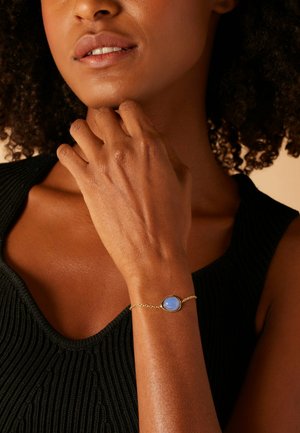 Gouden kettingarmband met een ovale blauwe steen, gezet in een gouden bezel, gedragen aan een hand met een gestructureerde donkere top. Glad huidtextuur zichtbaar.