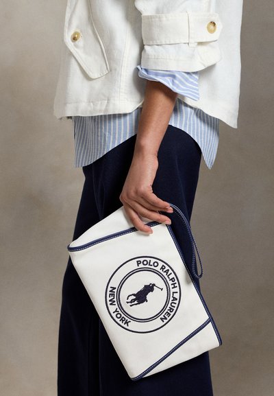 Χέρι που κρατά λευκό αξεσουάρ χειρός Polo Ralph Lauren με μπλε ραντάκι, φοράει λευκό μπουφάν, μπλε ριγωτό πουκάμισο και μπλε παντελόνι.