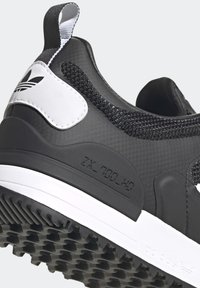Svart atletisk sko med texturerad tyg, syntetiska detaljer, vit sula och dragflik bak; har präglad logotyp och modellnummer.
