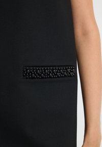 Schwarzes Kleidungsstück mit einer rechteckigen Tasche, die mit kleinen schwarzen Perlen verziert ist, gezeigt an der Seite einer Person, wobei ein Teil ihres Arms sichtbar ist.