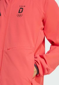 Koraalroze jas met rits, bedrukt met TEAM D en het Olympische logo. Heeft een gladde textuur, elastische manchetten en zijzakken voor functionaliteit.