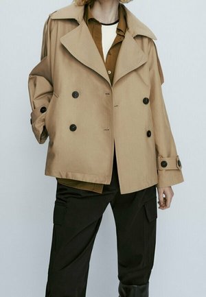 Persona che indossa un trench beige doppiopetto sopra una camicia marrone e pantaloni cargo neri, in piedi davanti a uno sfondo chiaro uniforme.