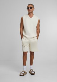 Vestă fără mâneci crem, pantaloni scurți asortați și sandale maro cu bretele. Șosete albe vizibile, design minimalist, textură moale.