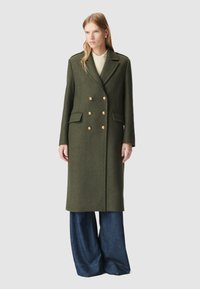 Cappotto verde in lana doppiopetto con bottoni dorati, due tasche frontali e un taglio sartoriale, abbinato a pantaloni in denim a gamba larga.