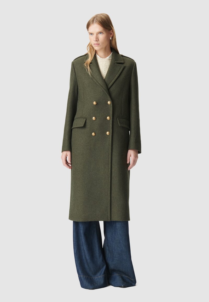 Cappotto verde in lana doppiopetto con bottoni dorati, due tasche frontali e un taglio sartoriale, abbinato a pantaloni in denim a gamba larga.