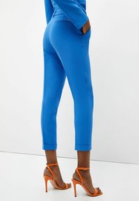 Blå skräddarsydda byxor med en slät textur, med uppvikta fållar. Åtföljs av orange högklackade sandaler, vilket framhäver kontrasten i färg.