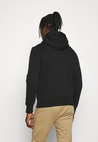 Black hoodie with a spacious hood, made of soft fabric. Features ribbed cuffs and hem. Worn with beige pants, viewed from the back.