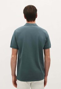 Paita polo dalam kain teal gelap, lengan pendek, dan kerah klasik. Permukaan bertekstur dengan hem lurus dan manset lengan yang dilipat.