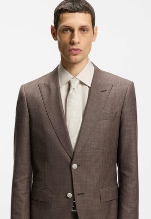 Mann trägt eine braune strukturierte Anzugjacke, ein beiges Hemd und eine gemusterte cremefarbene Krawatte, steht vor einem schlichten weißen Hintergrund.