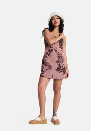 Robe rose sans manches avec des motifs de feuilles foncées, une silhouette ajustée et une légère texture. Associée à des sandales à plateforme blanches.