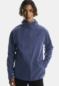Giacca impermeabile blu da uomo con cappuccio, zip frontale e logo. Caratterizzata da una texture liscia e un design aderente per l'uso attivo.