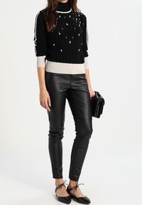 Schwarzer Strickpullover mit weißen Akzenten und Verzierungen, kombiniert mit eng anliegenden schwarzen Kunstlederhosen und schwarzen spitzen Flats.