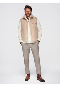 Gilet trapuntato beige, maglione crema a trecce, pantaloni grigi chiari e stivaletti anke in pelle marrone. Design semplice, colori neutri e stile aderente.