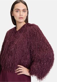 Veste courte texturée bourgogne avec des détails en franges brillantes, manches arrondies et coupe décontractée, portée avec un vêtement assorti en dessous.