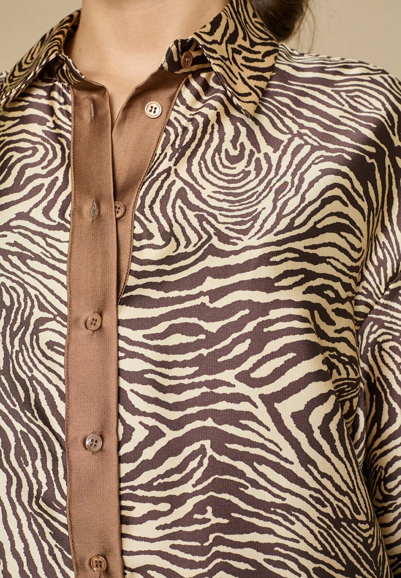 Brun og beige skjorte med zebra-print, lavet af glat satinmateriale, med knapper foran og en kontrasterende krave med brune knapper.