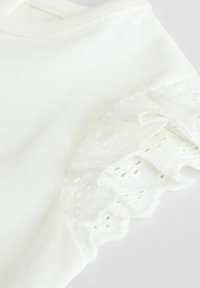 Chemise blanche en coton à manches courtes avec des détails de volants en dentelle aux manches, présentant de découpes florales délicates et une texture douce.