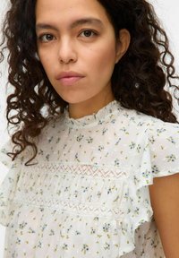 Blouse florale en tissu léger blanc avec de petites fleurs bleues, manches à volants, design plissé et accents en dentelle sur la poitrine.
