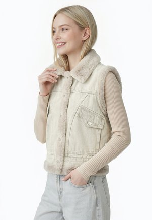Ung kvinde smiler, iført en beige ærmeløs vest med falsk pelsfor over en ribbet langærmet trøje og lyse denimjeans.