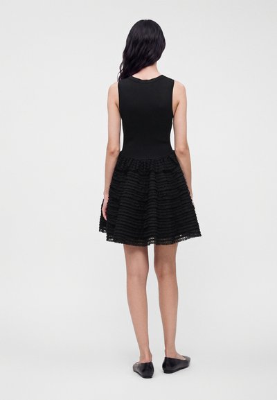 Femme aux longs cheveux foncés portant une robe noire ajustée sans manches avec une jupe évasée texturée et des chaussures plates noires, debout de dos.