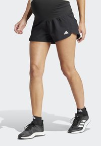 Pantalones cortos deportivos negros con una cintura fruncida, que cuentan con un logo de Adidas. Combinados con zapatillas negras y calcetines cortos, lo que muestra un estilo deportivo.