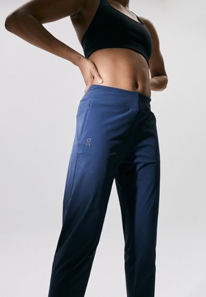 Personne en forme portant un soutien-gorge de sport noir et un pantalon de sport bleu marine avec une poche zippée, les mains reposant sur le bas du dos.