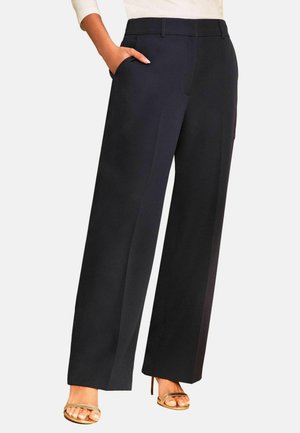 Vrouw draagt hoge taille zwarte wijde broek met hand in de zak en gouden open teen hoge hak sandalen.
