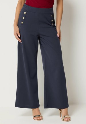 Mujer que lleva pantalones anchos de color azul marino con botones dorados en los bolsillos, combinados con sandalias doradas de tacón alto y una blusa roja.
