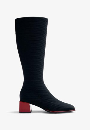 Botte noire hauteur genou avec une texture striée, bout pointu et un talon bloc rouge. Conçue avec une silhouette ajustée.
