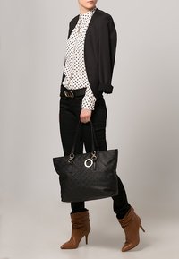 Grand sac cabas matelassé noir avec poignées doubles, élément décoratif circulaire en métal, associé à un chemisier à pois, un pantalon noir et des bottines à talons marron.