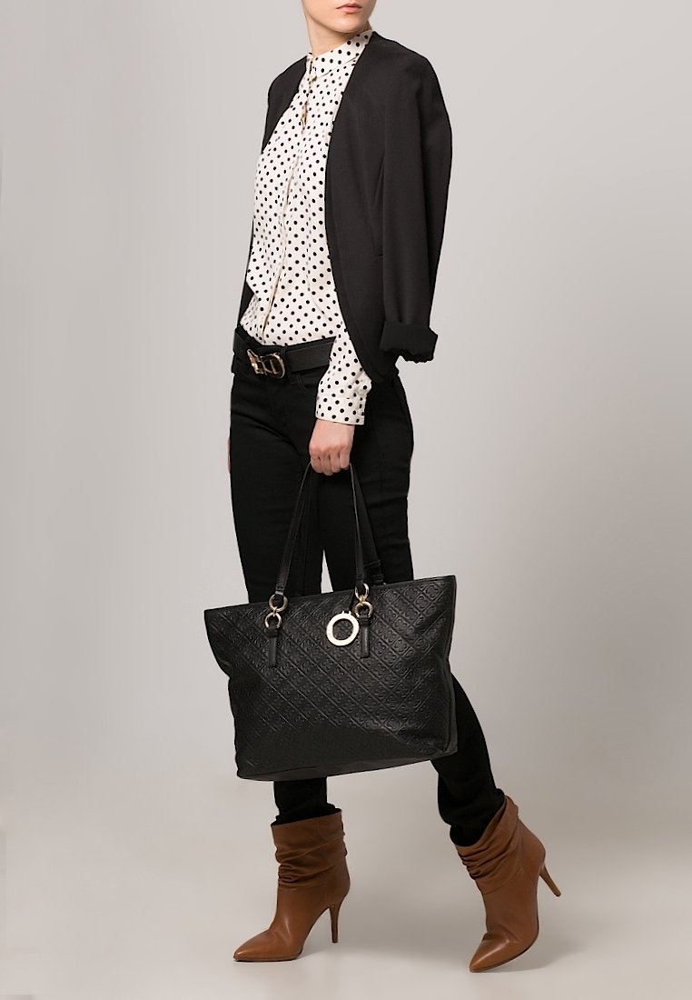 Grand sac cabas matelassé noir avec poignées doubles, élément décoratif circulaire en métal, associé à un chemisier à pois, un pantalon noir et des bottines à talons marron.