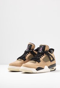 Paire de baskets Air Jordan 4 beige et noires avec lacets noirs, panneaux en mesh et semelles blanches sur une surface blanche.