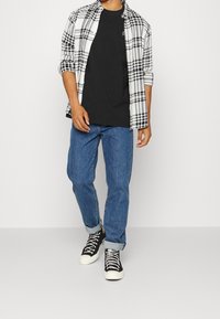 Czarna koszulka pod białą i czarną koszulą w kratę, zestawiona z niebieskimi dżinsami i czarnymi trampkami za kostkę z białymi podeszwami.