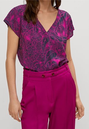 Kvinde iført magenta højtaljede bukser og en lilla kortærmet bluse med blomsterprint og V-hals, stående foran en ensfarvet baggrund.