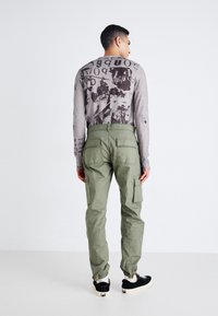 Pantalones cargo verdes con puños elásticos, dos grandes bolsillos laterales y bolsillos traseros. El modelo lleva una camiseta gráfica gris de manga larga y zapatillas deportivas.