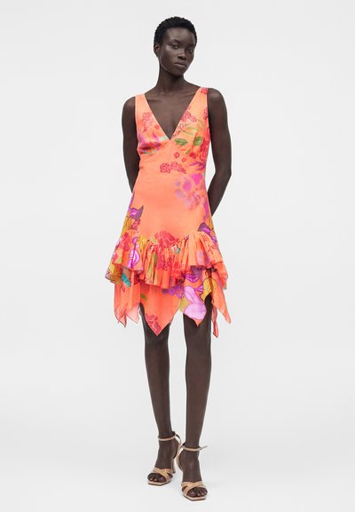 Γυναίκα με πορτοκαλί φόρεμα χωρίς μανίκια με floral σχέδιο και ασύμμετρο βολάν στο τελείωμα, και μπεζ ψηλοτάκουνα σανδάλια με λουριά σε λευκό φόντο.