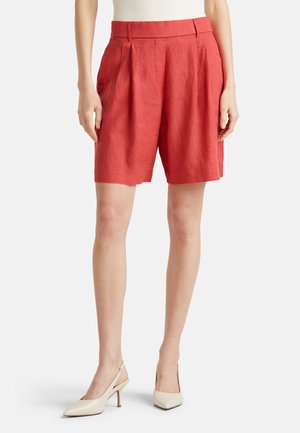 Bermudas de lino rojas con pliegues, una cinturilla alta, bolsillos laterales y un ajuste relajado, combinadas con zapatos de tacón alto en color crema.