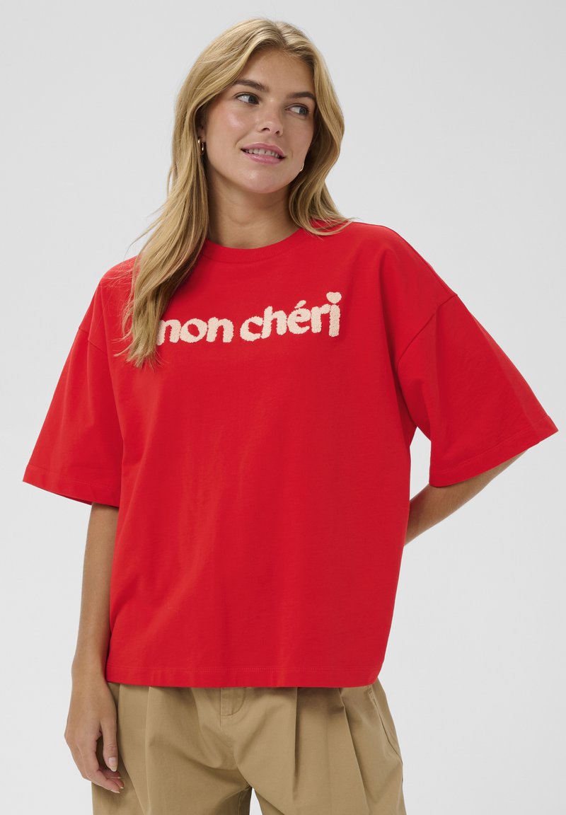 Sarkana kokvilnas T-krekls ar īsām piedurknēm un vaļīgu piegriezumu ar reljefām baltām burtiem priekšpusē, uzraksts “mon chéri”.