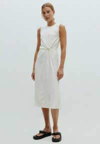Witte mouwloze jurk met een geknoopte detail bij de taille, met een gladde textuur en een midi-lengte. Zwarte plateausandalen maken de look compleet.
