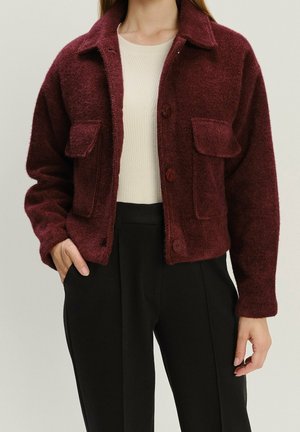 Vrouw met een getextureerd bordeaux jasje met knopen en voorzakken, een crèmekleurige geribde trui en een zwarte broek met hoge taille.