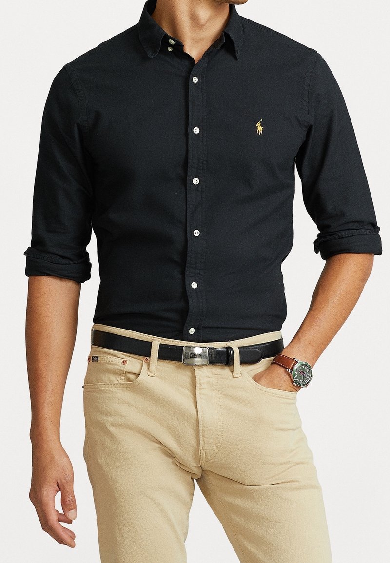 Chemise noire à manches retroussées, ornée d'un petit logo jaune sur la poitrine. Assortie à un pantalon beige clair et une ceinture noire.