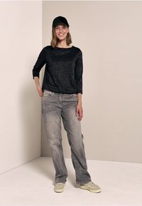 Schwarzes glitzerndes Langarmoberteil, hellgraue gerade Jeans und hellen Sneaker, dazu eine schwarze Kappe. Das Model steht vor einem neutralen Hintergrund.
