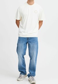 T-shirt crème à manches courtes avec détail de logo circulaire sur la poitrine, associé à un jean bleu clair à coupe ample et des baskets grises.