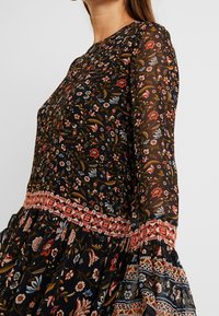 Mujer vistiendo un vestido floral negro transparente con flores rojas, azules y blancas, y un detalle de cintura y mangas en rojo con patrón.