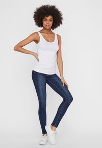 Débardeur blanc et jean skinny bleu foncé associés à des baskets blanches ; le haut est sans manches avec un col arrondi, le tissu semble lisse.